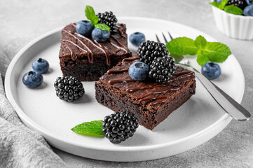 Chocolate brownies with chocolate glaze, fresh berries and mint leaves on top on a white plate on a...