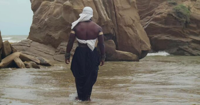 A cosplaying male with dark skin and muscles on the sand with a cliff in the background