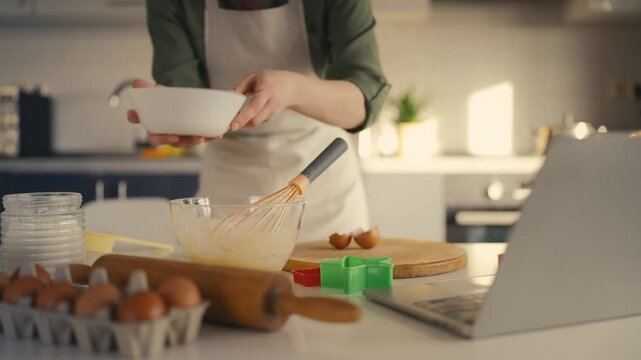 Woman watching cooking video on laptop, stirring ingredients in kitchen bowl
