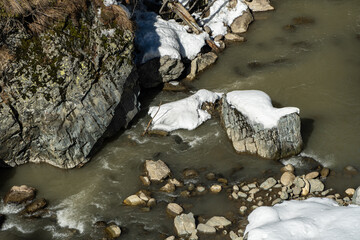 Winter scene of rocky riverbank with snow-covered rocks and calm, flowing water, capturing the tranquility of nature in cold season. Karachay-Cherkessia. Dombay 2025. Nature concept for design.