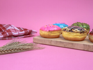 Colorful assorted donuts on a wooden board with pink background, wheat stalks, and a red napkin