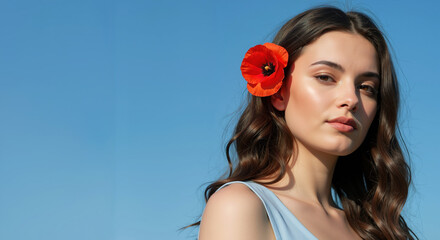 Naklejka premium Woman with wavy brown hair and red poppy flower behind ear against blue background. Natural floral beauty concept for summer fashion, cosmetic advertising and seasonal accessories marketing