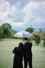 A Muslim couple stands close under a clear umbrella in a lush green field, facing away from the camera. Captured from behind, the moment reflects love, peace, and togetherness on a cloudy day