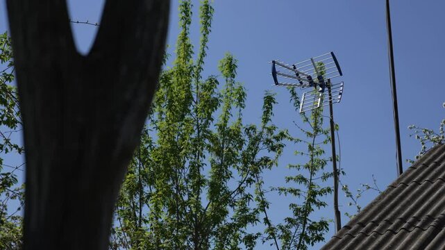 Outdoor passive television decimeter antenna on a long pole among trees against a blue sky near a one-story private house with a slate roof. Analog and digital radio signal receiver in remote areas.