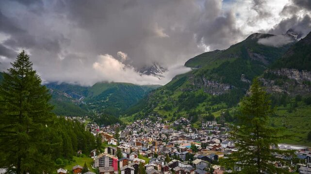 Timelapse of heavy clouds moving above the alpine town of Zermatt, surrounded by lush green hills and mountain peaks of Swiss Alps in Switzerland. 4K UHD video.