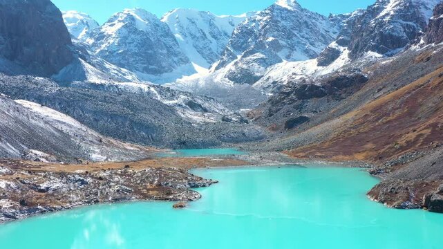 Turquoise waters of lake surrounded by mountain range. Snow capped rock peaks and glaciers on clear sunny day. Aerial view, flying back. Crystal clean water. Untouched nature. Hiking in the mountains.