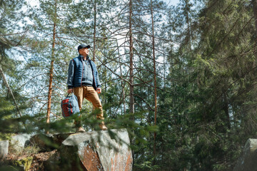 Smiling man with glasses and backpack hiking in a beautiful pine forest. Standing on mossy rocks, looking up. Outdoor adventure, positive emotion.