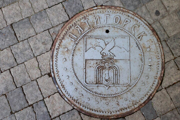 Round Metal Manhole Cover On A Cobblestone Pavement, Featuring The Coat Of Arms Of Pyatigorsk And The City Name In Cyrillic. Urban Utility Detail, Russia.