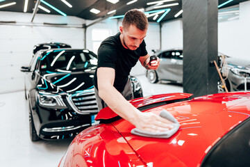 Professional car detailer using an orbital polisher to polish the hood of a red car, ensuring a...