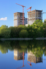 Obraz premium Two High-Rise Buildings Under Construction With Red Tower Cranes, Perfectly Reflected In The Calm Blue Water Of A Lake Or Pond. Green Trees Line The Shore.