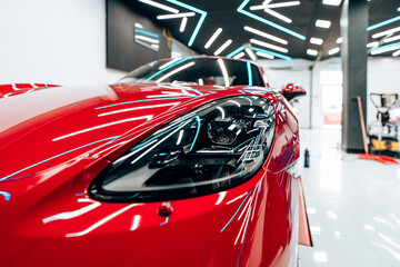 Neon lights reflecting off the polished hood and gleaming headlight of a vibrant red sports car, receiving meticulous detailing in a modern, brightly lit workshop environment. 