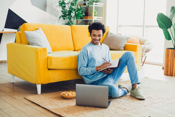 Young man relaxing on the floor at home wearing a blue sweatshirt and jeans writing in a notebook while using a laptop.