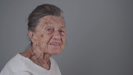 Senior woman 90 years old smiles, laughs and shows a denture on a gray background. Elderly female with deep wrinkles and gray hair happy in a good mood studio portrait looking at camera.