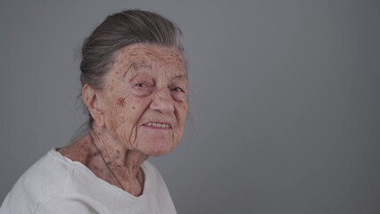 Senior woman 90 years old smiles, laughs and shows a denture on a gray background. Elderly female with deep wrinkles and gray hair happy in a good mood studio portrait looking at camera.