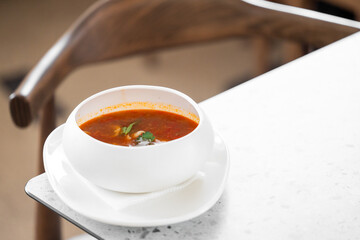 A white bowl of tomato soup garnished with fresh herbs sits on a white plate with a napkin, placed...