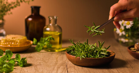 A hand is using tongs to pick up rosemary leaves from a wooden plate below. The rosemary leaves are green and have small needle-like leaves. A few basil leaves are scattered on the wooden table.