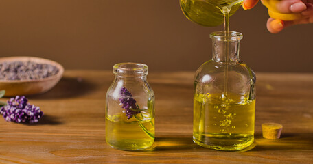 The action of pouring essential oil into a glass bottle causes bubbles to rise in. There is a sprig of lavender in the bottle. Behind it is a dish containing lavender seeds. Brown background.
