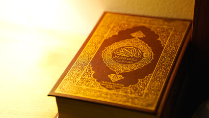 Closed Quran book with ornate golden Arabic design on brown cover resting on table with warm soft lighting reflecting Islamic spirituality