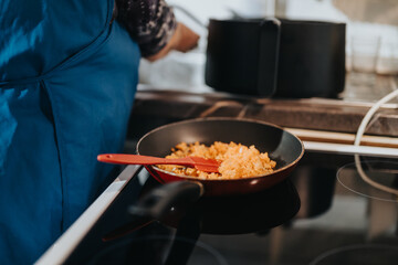 An individual preparing a dish in a cozy kitchen setting, focused on a frying pan with an orange utensil. Warm lighting enhances the culinary ambiance.