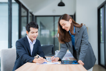 Two asian businesspeople are smiling and working together, analyzing financial charts and graphs at a desk in a modern office, using pens and a laptop