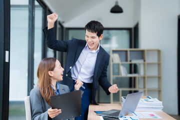 Two asian businesspeople celebrating success while working together on laptop computer in modern office, business team celebrating successful contract, business achievement concept