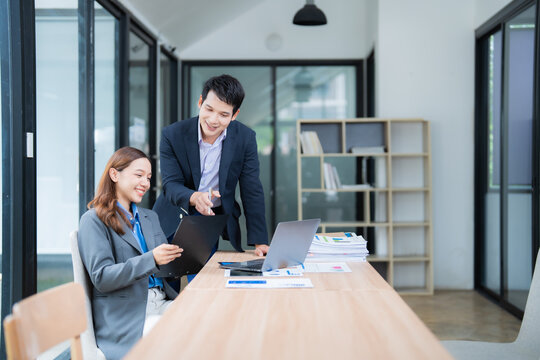 Two asian businesspeople are discussing and working together, using a laptop and analyzing paperwork with charts and graphs, in a modern office