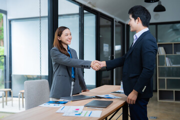 Two Asian businesspeople shaking hands in a modern office, smiling at each other after successfully closing a deal. Collaboration and teamwork evident in their confident expressions