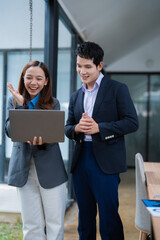 Two asian business people wearing suits are standing in the office, looking at a laptop and having a video conference, smiling and gesturing