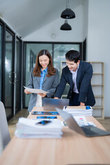 Two Asian businesspeople collaborating in an office, analyzing financial data while working on a laptop and reviewing important documents for effective decision-making