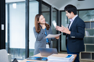 Two Asian businesspeople standing near a desk, discussing and collaborating while holding documents and smiling in a bright, modern office environment filled with natural light