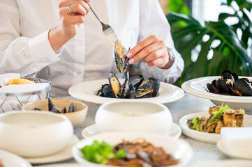 person in a white shirt enjoys eating mussels at a restaurant table set with seafood, bread, and salad. The close-up captures a fine dining experience with fresh gourmet cuisine.