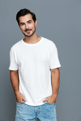 Smiling young man wearing a white t-shirt stands confidently against a gray background, looking directly at the camera with a friendly expression.