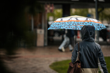 A woman dressed in a two-tone raincoat and jeans walks on a wet brick path holding a vibrant, patterned umbrella, with spring greenery and urban buildings in the background.