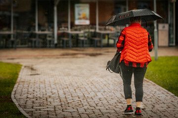 A woman wearing a red plaid shirt and vest walks on a wet sidewalk under a gray umbrella during a rainy day, carrying a quilted black handbag.