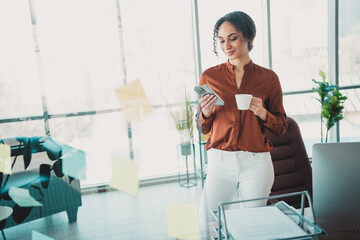 Young professional businesswoman enjoying coffee while multitasking with her smartphone in a modern...