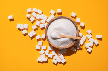 White granulated sugar in wooden bowl and scattered sugar cubes on yellow background top view....