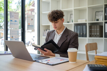 Young businessman reviewing notes while working on a laptop at a modern office desk.