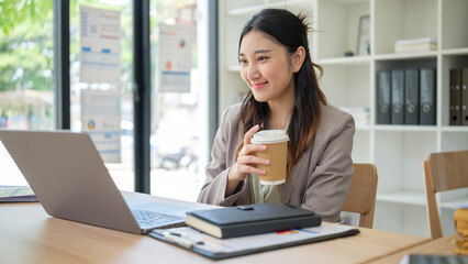 Fototapeta premium Smiling businesswoman holding a coffee cup and looking at her laptop, taking a short break during work.