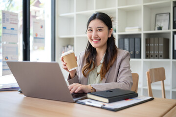 Confident young businesswoman working on her laptop while enjoying coffee in a modern office environment.