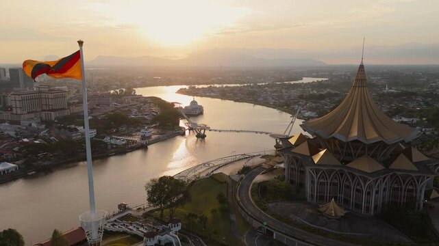 Stunning aerial view of Kuching at sunset, featuring the iconic Sarawak State Legislative Assembly building, DUN, the Darul Hana Bridge, and the Sarawak River. The Sarawak flag waves proudly.