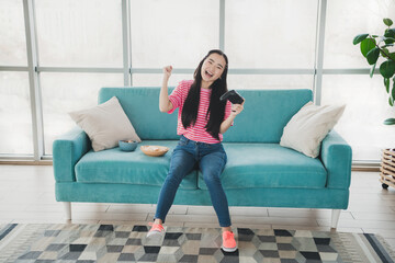 Obraz premium Young woman smiling while gaming on a turquoise couch in a modern living room with snacks and natural daylight