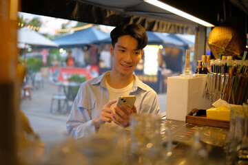 Young man smiling while using his smartphone at a vibrant outdoor food stall.