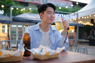 Young man enjoying beer and street food at night market with festive lights and food stalls in the background.