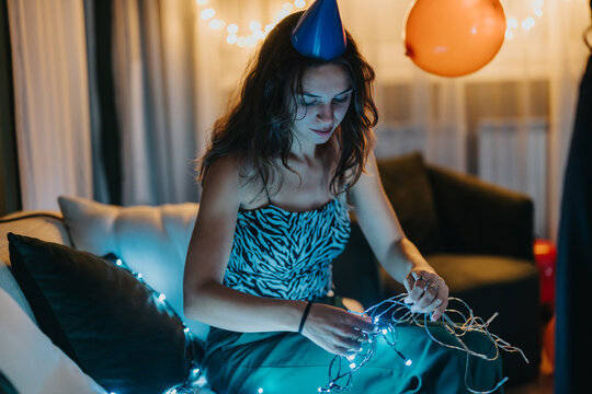 A young woman wearing a party hat prepares string lights while sitting on a couch indoors. The ambiance is decorated with festive balloons and illuminated by fairy lights, symbolizing celebration and