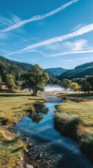 Obraz premium Serene valley landscape with a clear stream, green meadows, trees, distant hills, and a blue sky streaked with clouds.