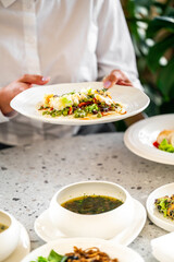 person in a white shirt holds a plate of colorful food with vegetables and seafood. meal is set on a dining table with bowls of soup and other dishes, suggesting fresh and gourmet culinary experience