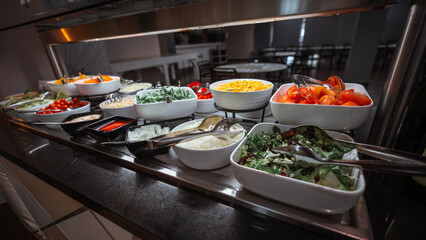 Colorful self-service salad buffet with a variety of fresh vegetables including tomatoes, corn, onions, lettuce, arugula, cherry tomatoes, and sauces, displayed in white bowls under warm lighting