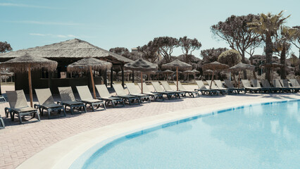 Serene resort swimming pool surrounded by empty lounge chairs and thatched umbrellas, with a tropical tiki bar in the background and palm trees under a clear blue sky, perfect vacation scene