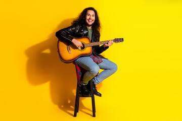 Fototapeta premium Young fashionable man seated on stool playing acoustic guitar against bright yellow studio background