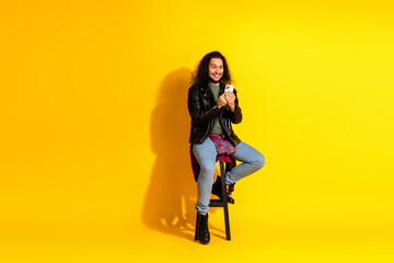 Young stylish man in a leather jacket sitting on stool holding a smartphone against yellow background.